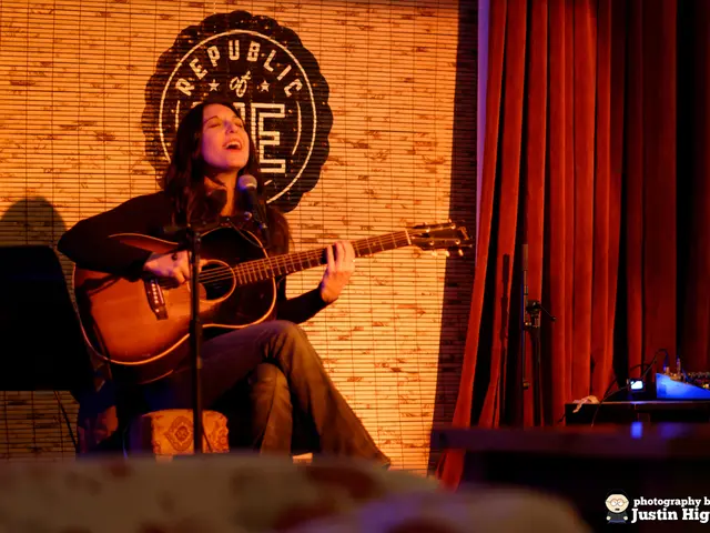 The picture consists of one person sitting on the chair and playing a guitar and singing in front...